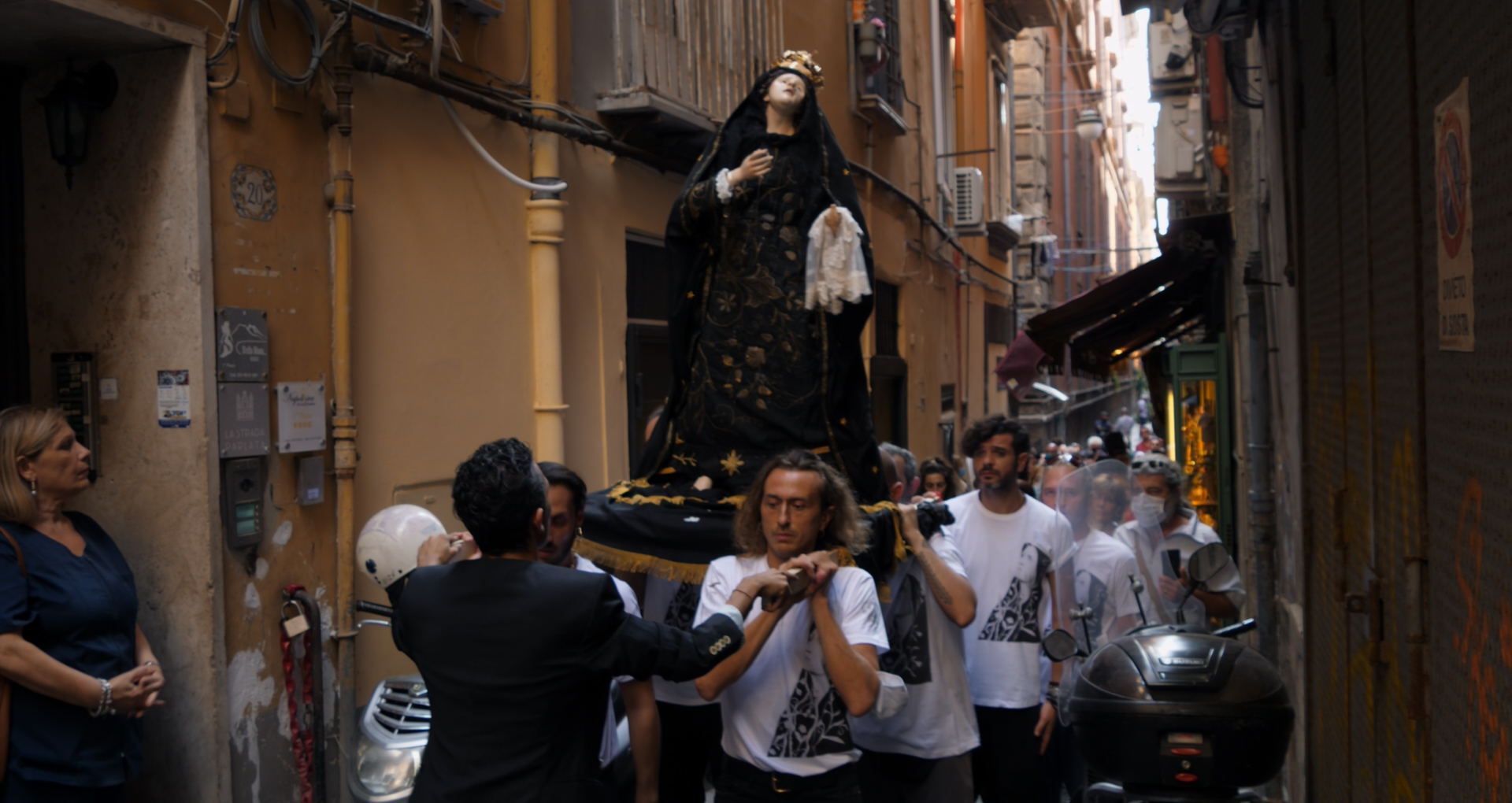 A frame from the film “The Gospel According to Ciretta,” showing a narrow street in Naples through which a procession of men in white T-shirts is passing, carrying a statue of Mary.