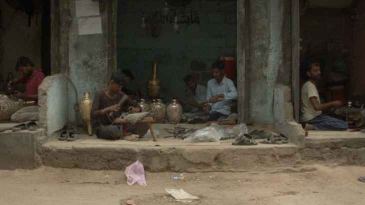 Kadr z filmu "A Song of Humble Beauty" przedstawiający uliczkę w Karachi, przy której grupka mężczyzn tworzy rękodzieło. Wokół nich znajdują się różne zdobione wazy.