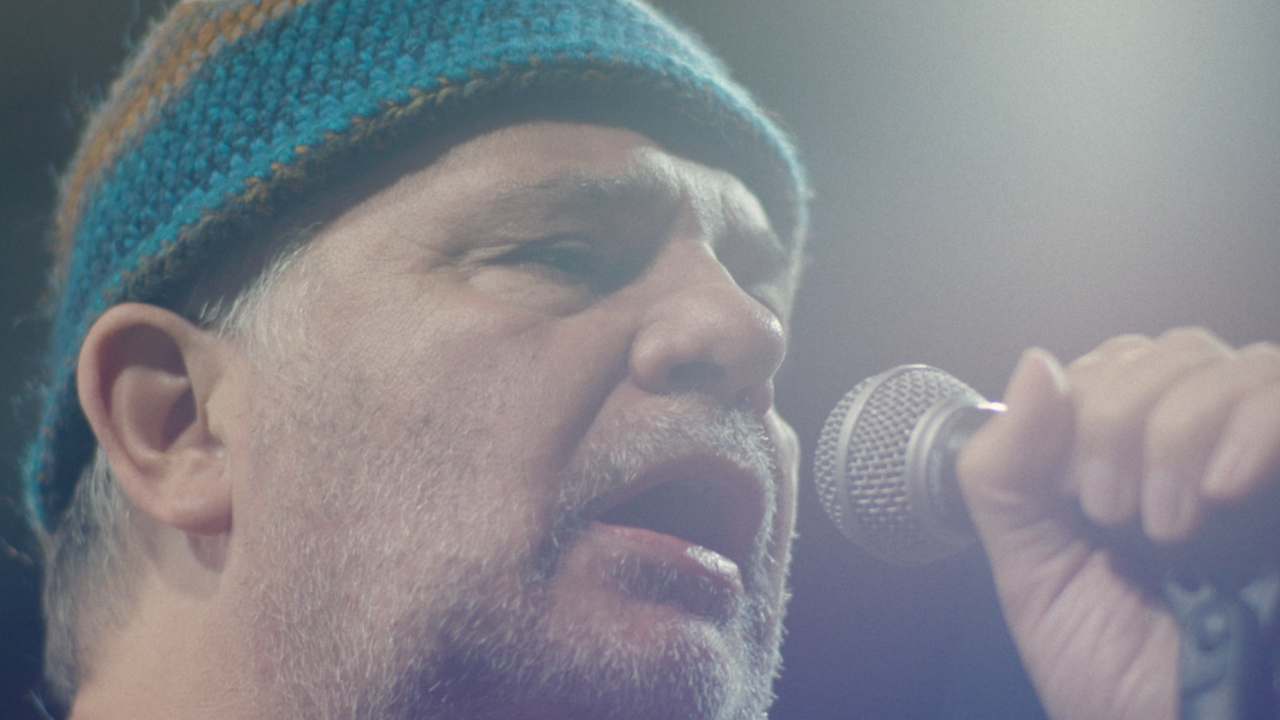 Kadr z filmu "Pieśń wieloryba" przedstawiający przybliżenie na twarz mężczyzny w średnim wieku w niebiesko-żółtej czapce na głowie oraz z zarostem na twarzy. Mężczyzna przemawia do mikrofonu trzymanego w dłoni.