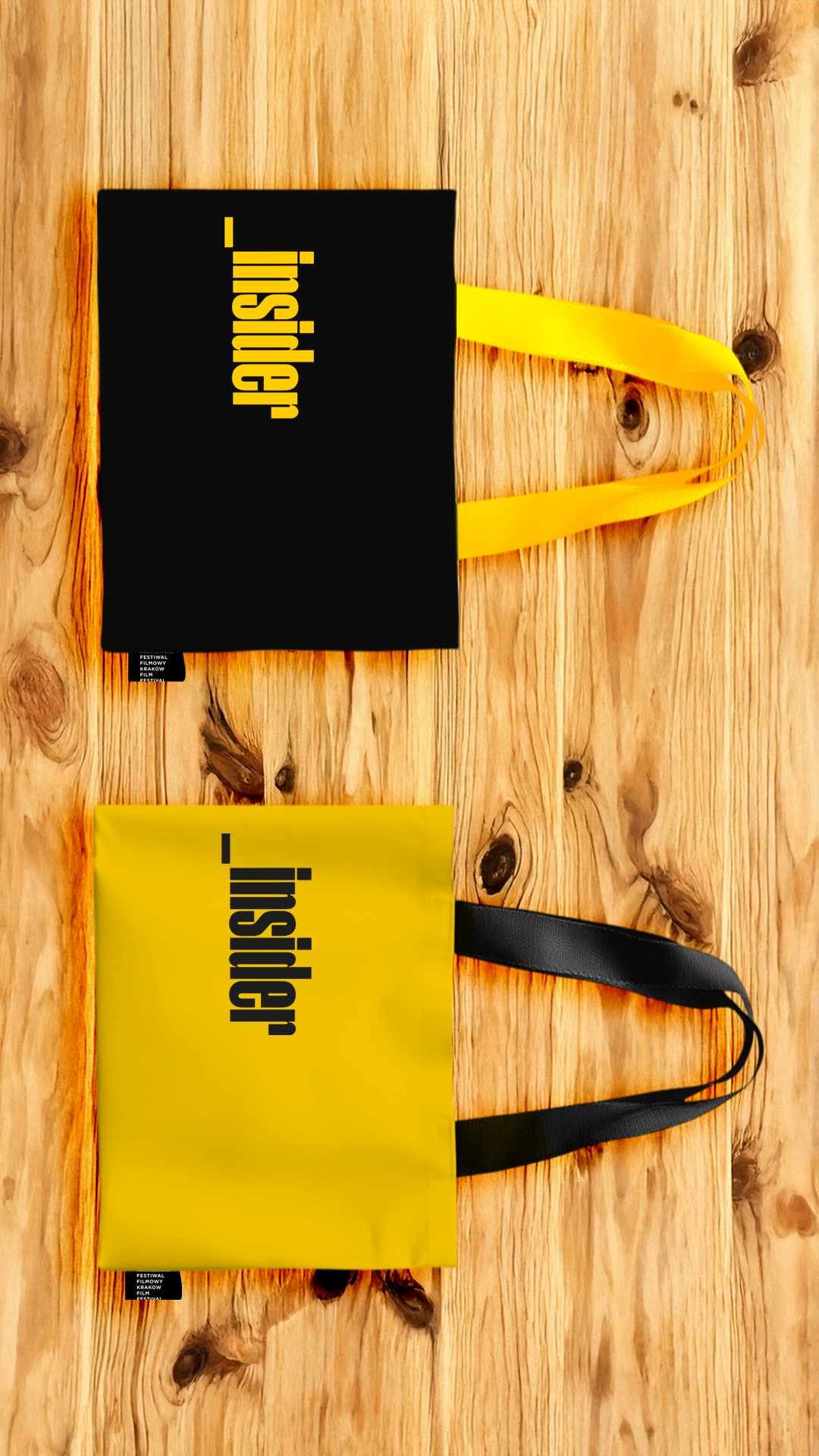 Dwie materiałowe torby _insider leżące obok siebie na jasnym, drewnianym blacie: czarna z żółtym uchem i napisem oraz żółta z czarnym uchem i napisem